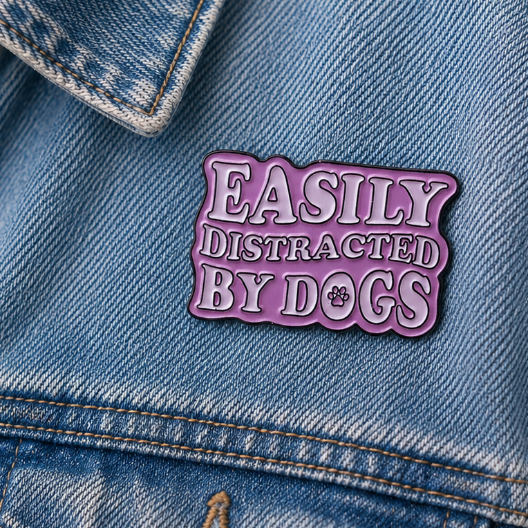 A die-cut enamel pin with the phrase 'EASILY DISTRACATED BY DOGS' in white raised lettering with a dark outline, set against a light Pink background with a small paw print detail.