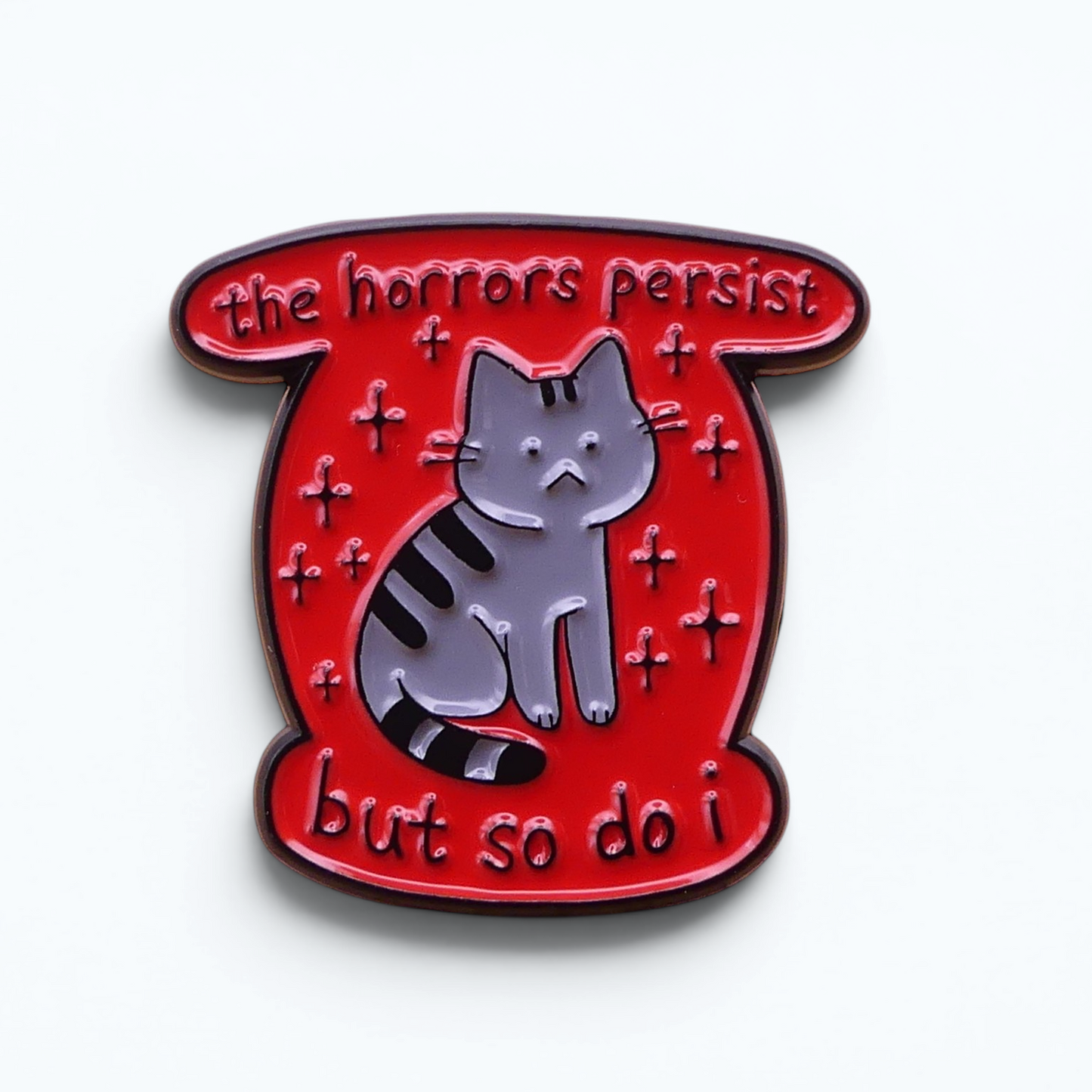 A red enamel pin shaped like a scroll, featuring a grey tabby cat with black stripes and the text 'the horrors persist but so do I.
