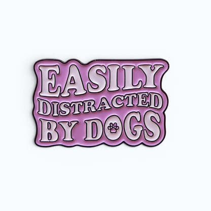 A die-cut enamel pin with the phrase 'EASILY DISTRACATED BY DOGS' in white raised lettering with a dark outline, set against a light Pink background with a small paw print detail.