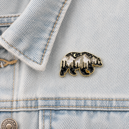 Close-up black enamel bear pin with stars, moon, and pine trees in gold trim