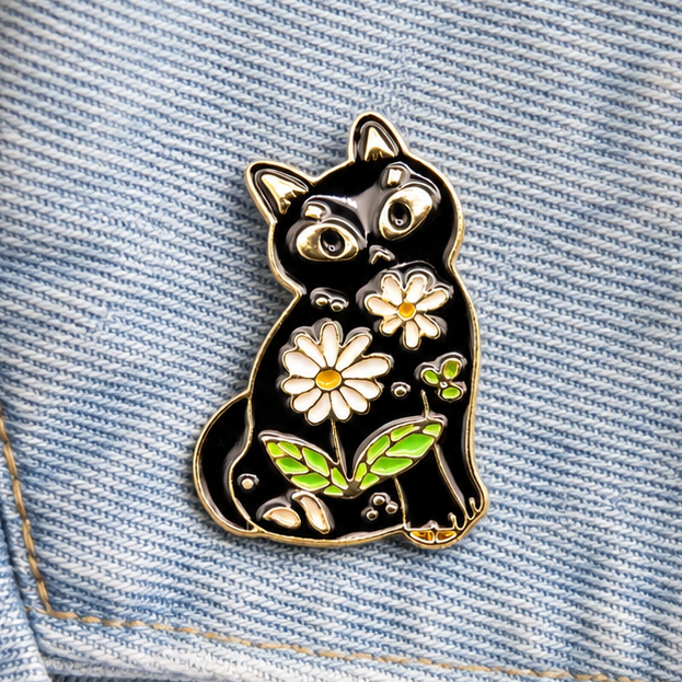 Black cat enamel pin with daisies and green leaves on light-blue denim fabric.