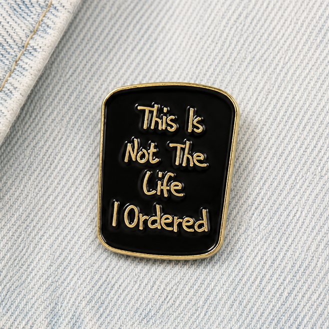 Square black enamel quote pin badge with gold border on light denim fabric