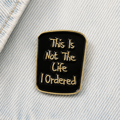 Square black enamel quote pin badge with gold border on light denim fabric