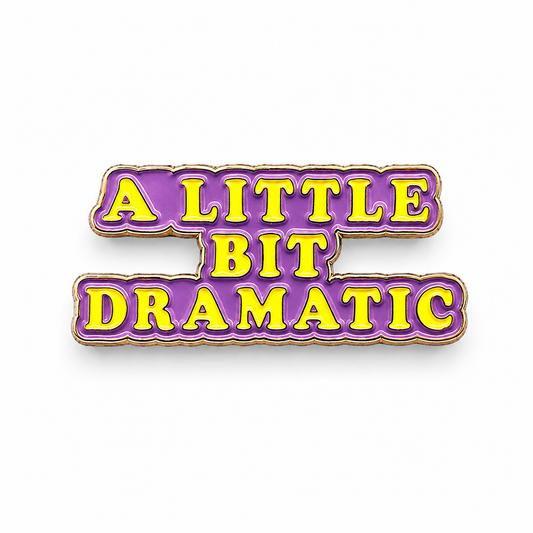 Purple enamel text badge pin reading “A LITTLE BIT DRAMATIC” with gold border.