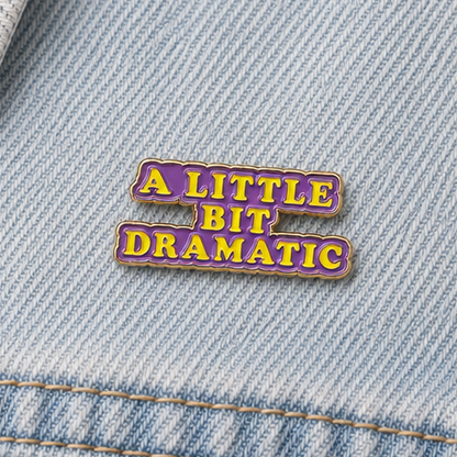 Purple enamel pin with gold outline on blue denim, “A LITTLE BIT DRAMATIC” text.