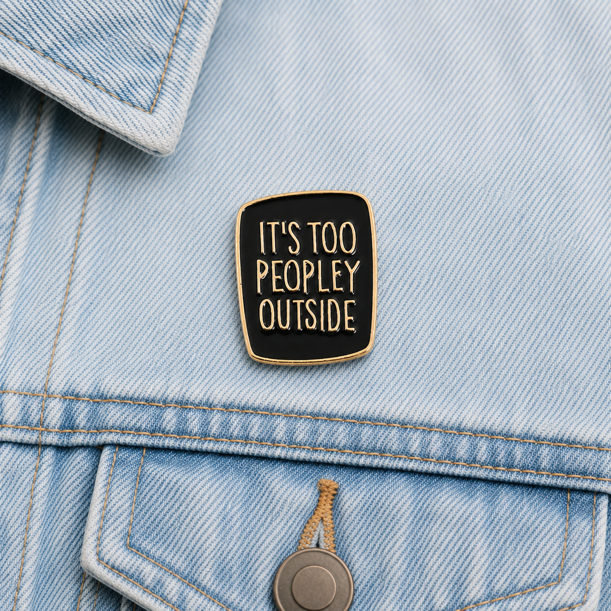 Enamel pin badge on light blue denim: black square with gold lettering “It’s too peopley outside”.