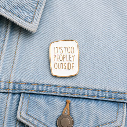 Enamel pin badge on light blue denim: white square with gold border and “It’s too peopley outside” text.