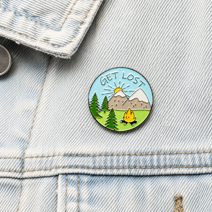 Round enamel pin badge on light denim: mountains, trees, campfire.