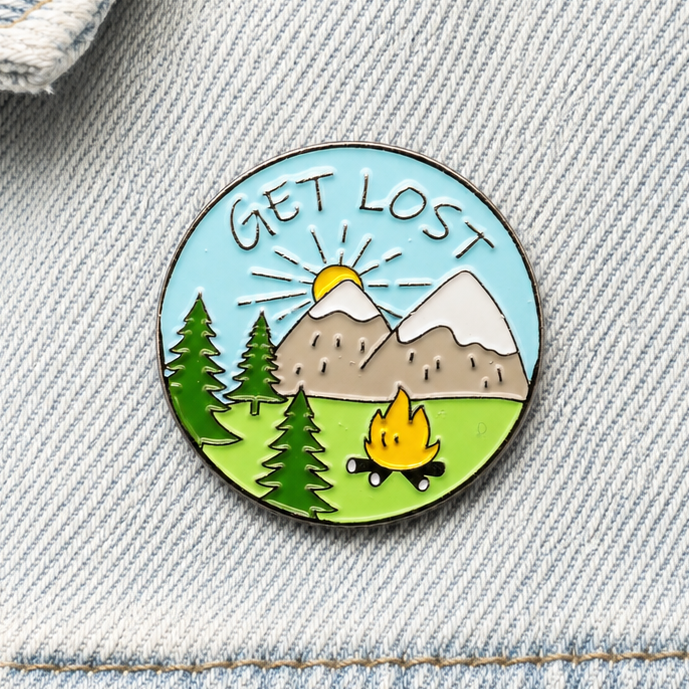 Get Lost enamel pin badge close-up with mountains, trees and campfire scene.