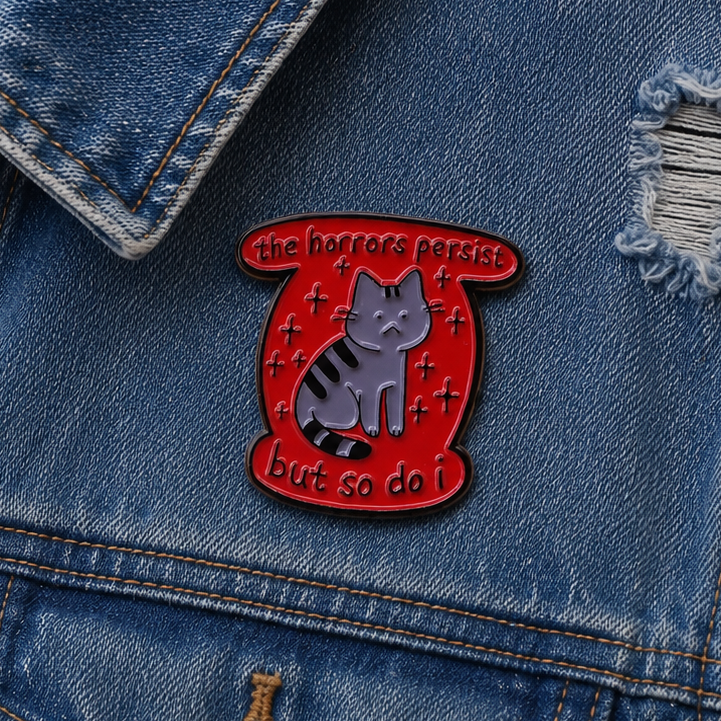 A red enamel pin shaped like a scroll, featuring a grey tabby cat with black stripes and the text 'the horrors persist but so do I.