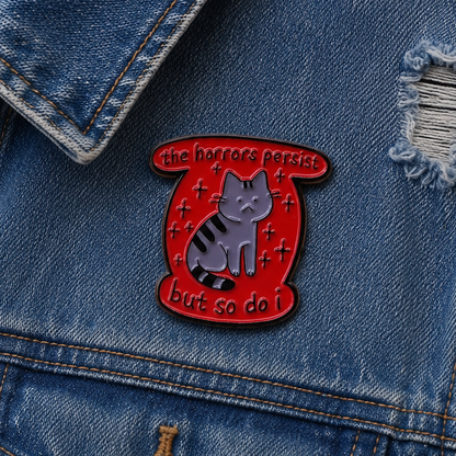 A red enamel pin shaped like a scroll, featuring a grey tabby cat with black stripes and the text 'the horrors persist but so do I.