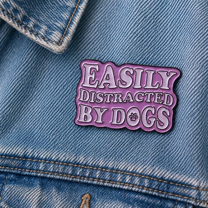 A die-cut enamel pin with the phrase 'EASILY DISTRACATED BY DOGS' in white raised lettering with a dark outline, set against a light Pink background with a small paw print detail.