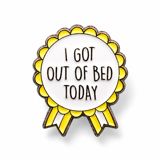 A circular enamel pin shaped like a ribbon badge with yellow scalloped edges, black metal outlines, and the text 'I GOT OUT OF BED TODAY' on a white center, shown on a light gray background.