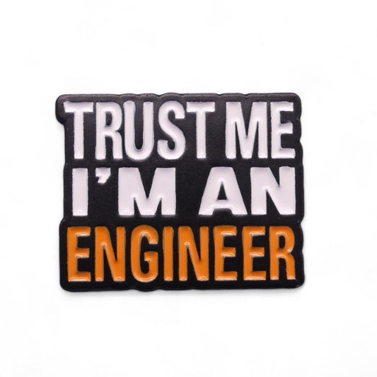 Black enamel pin badge with white and orange text saying Trust Me I'm An Engineer.